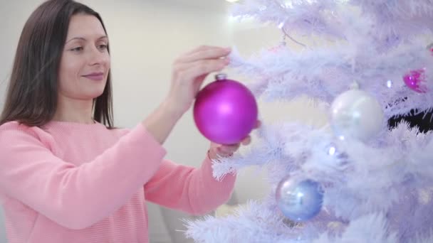 Aantrekkelijke zakenvrouw versiert nieuwjaarsboom met roze glazen kerstballen, op zoek naar blij met het proces. — Stockvideo