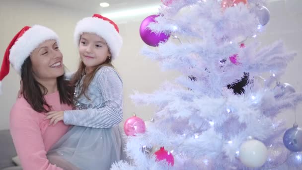 Moeder houdt haar dochter, staan in de buurt van kerstboom, schattig meisje wijst naar een van roze kerstballen op de sparren tak. — Stockvideo