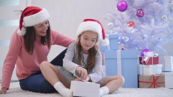 Mamma och dotter inslagning presenter, sitter på mattan nära julgranen i nyårstid. — Stockvideo