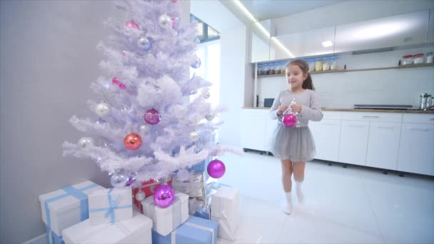 Hübsches kleines Mädchen kommt an den Weihnachtsbaum auf der Vorderseite und legt eine Glaskugel auf einen der Zweige. — Stockvideo