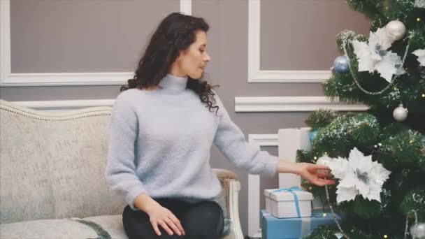 Pretty young woman is sitting on a sofa near Christmas tree, enjoying holiday spirit. — Stock Video