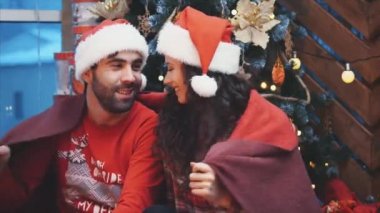 Mutlu adam ve kadın Noel ağacının yanında oturuyor, kırmızı battaniyeye sarılıp burunlarına dokunuyorlar..