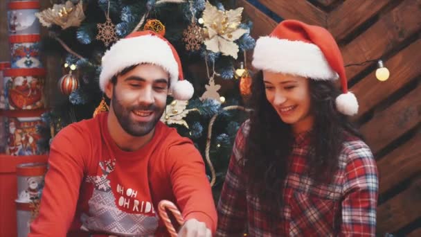 Two sweet people in love are sitting side by side and holding candy canes, so that they form a heart, over wooden planks background. — Stock Video