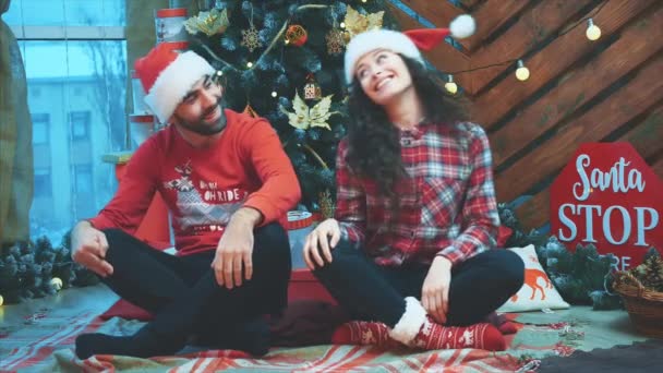 Fresco divertente giocoso partner indossato in maglia vestiti rossi e cappelli di Babbo Natale sono scherzare in giro, ballare, seduto in posizione foto . — Video Stock