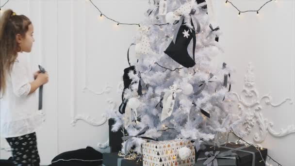 Kleines brünettes Mädchen schmückt einen Weihnachtsbaum mit gestreiften Schleifen, tanzt und lächelt und genießt diesen Beruf. — Stockvideo