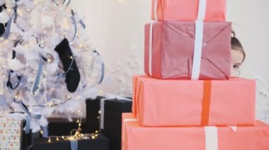 Güzel küçük kız Noel hediyesi kutularıyla oynuyor, bir piramit inşa ediyor ve arkasından, bir tarafından, diğer tarafından dışarı bakıyor..