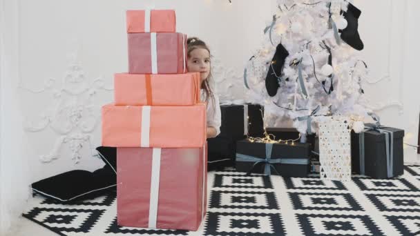 Bella bambina che gioca con le scatole regalo di Natale, costruisce una piramide e guarda fuori da dietro, da un lato, poi da un altro . — Video Stock