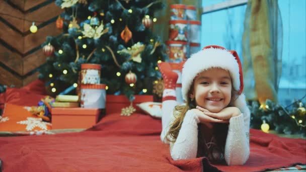 Santa Claus niña acostada en la manta roja bajo el árbol de Navidad, moviendo sus piernas en calcetines rojos, mirando a la cámara feliz y complacido . — Vídeo de stock