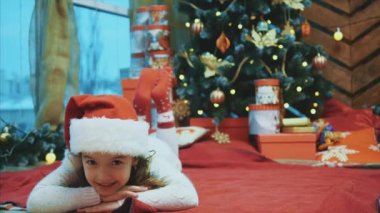 Komik bir video. Kırmızı Noel Baba şapkalı şirin bir cüceye benzeyen, köknar ağacının altındaki kırmızı battaniyeye uzanmış komik surat ifadeleri sergiliyor..