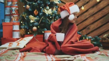 Noel Baba şapkalı uykulu çocuk, Noel ağacının yanında kırmızı battaniyeye sarılı, bir fincan kakaolu, keyifli ve memnun görünüyor..