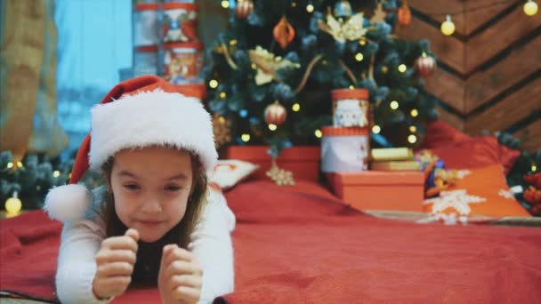 Rolig video där liten unge ligger på röd filt under gran-träd, gör komiska ansiktsuttryck, ser ut som söt tomte, klädd i röd tomte hatt. — Stockvideo