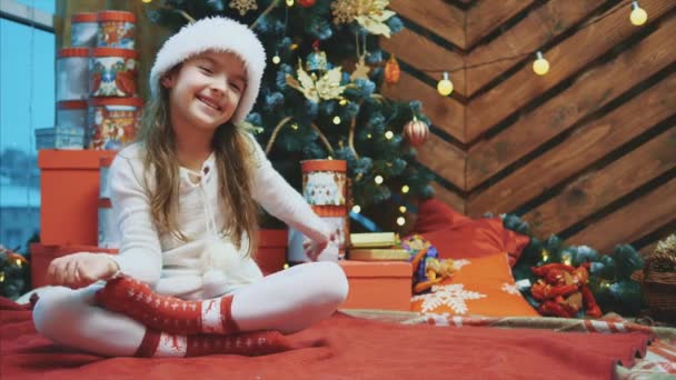 4kビデオの若いです女性身に着けているクリスマスの帽子手を振って、招待誰もが彼女と一緒に木の背景. — ストック動画
