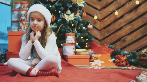 Enttäuschtes kleines Mädchen im Weihnachtskostüm sitzt in Lotos-Position vor hölzernem Weihnachtshintergrund und runzelt die Stirn. — Stockvideo