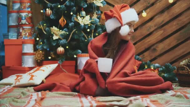 Criança sonolenta em chapéu de Papai Noel, envolto em cobertor vermelho perto da árvore de Natal, com uma xícara de cacau, olhando encantado e satisfeito . — Vídeo de Stock