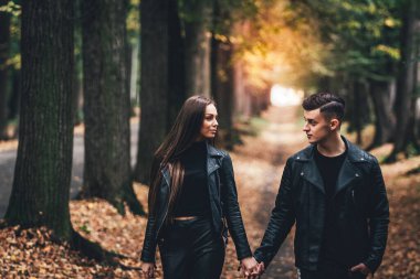 Birbirlerine aşık çiftin dramatik bir fotoğrafı. Siyah deri ceketli, el ele tutuşmuş ve sonbahar parkında geziniyorlar..
