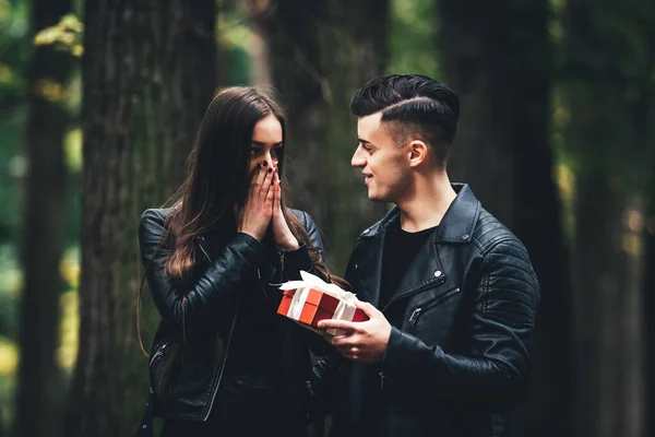 Handsome brunette man gives her girlfriend red present box, in autumn park. She looks astonished covering mouth by hands. — Stock Photo, Image