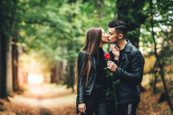 Leidenschaftliche junge Mann und Frau küssen sich im herbstlichen Park und halten eine rote Rose zusammen. — Stockfoto