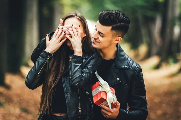 Schöner brünetter Mann bedeckt Frauenaugen mit der Hand und schenkt ihr eine rote Geschenkschachtel im Herbstpark. — Stockfoto