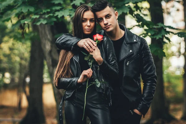 Superbe couple à la mode en noir posant à l'extérieur avec rose rouge dans les mains, regardant sans émotion et confiant . — Photo