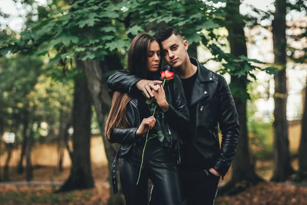 Gorgeous fashionable couple in black posing outside with red rose in hands, looking unemotional and confident.