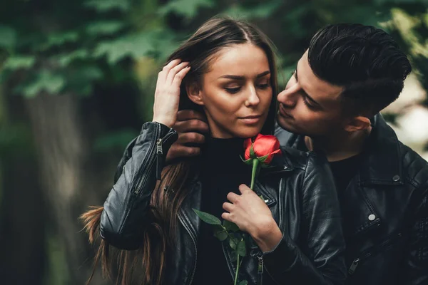 Morena homem abraçando sua namorada por trás com intenção de beijar, enquanto ela segura uma bela rosa . — Fotografia de Stock