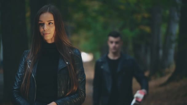 Menina morena em casaco de couro preto está esperando por seu namorado. Menino no olhar preto total está dando a sua linda namorada uma caixa de presente embrulhado vermelho. Fundo desfocado. Espaço para cópia. 4K . — Vídeo de Stock