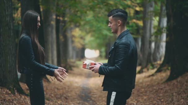 Jongen in zwart leren jas geeft zijn lieftallige vriendin een rode geschenkdoos. Wazige achtergrond. Langzame beweging. Begrepen, ruimte. 4K. — Stockvideo