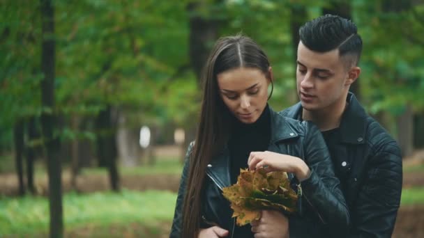 Garçon en manteau de cuir noir étreint sa charmante petite amie. Il tient un tas de feuilles d'automne, et la fille en prend et souffle à son tour. Fond flou. Ferme là. Espace de copie. 4K . — Video