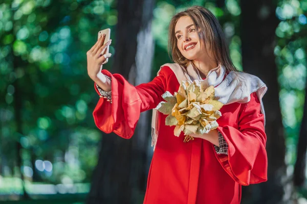 Wanita kaukasia yang menang mengambil selfie dengan seikat daun di luar ruangan di taman musim gugur. — Stok Foto