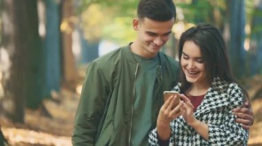 Genç, güzel bir çift patikada duruyor. Çift telefona bakıyor, sonbaharın bulanık arka planında. Kapatın. Uzayı kopyala 4K.