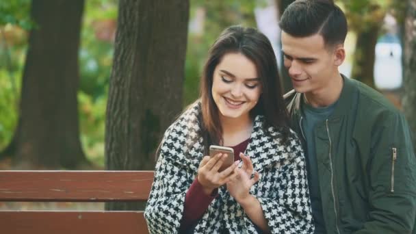 Paar kijkt samen naar smartphone, zittend op een bankje in het park. 4K. — Stockvideo