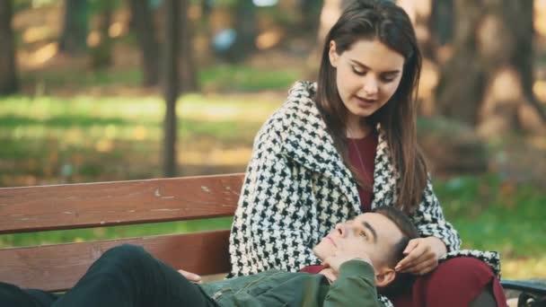 Jovem casal feliz está relaxando no parque em um banco. O tipo está encostado ao colo da namorada. Fecha. 4K — Vídeo de Stock