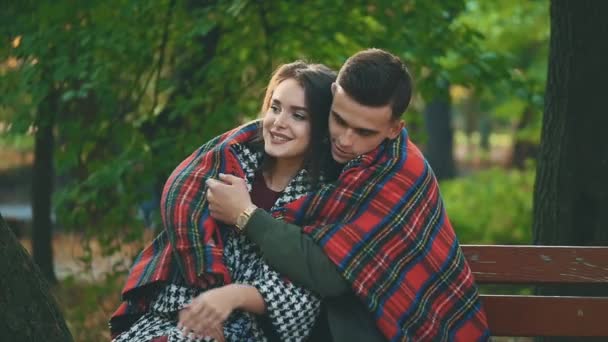 Jonge mooie paar knuffels met gezellige deken, zittend op een bank, op wazig park achtergrond. 4K. — Stockvideo