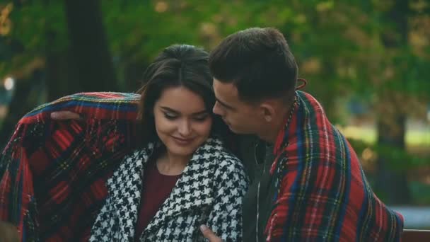 Lento. Joven pareja bonita abrazos con manta acogedora, sentado en un banco, sobre fondo borroso parque. 4K . — Vídeos de Stock