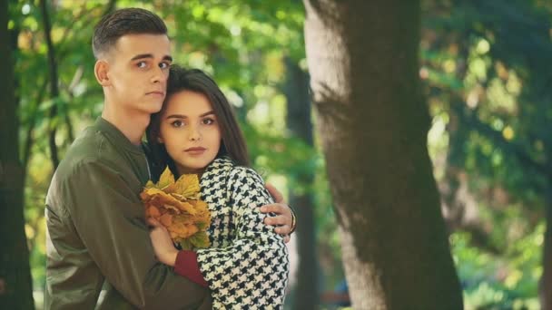 Lento. El chico está abrazando a su encantadora novia. Ella sostiene un montón de hojas de otoño. Fondo borroso. De cerca. Copiar espacio. 4K . — Vídeos de Stock