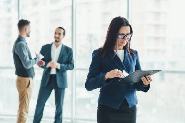 Önde duran genç bayan asistan imzalanmaya hazır belgelerle, arka planda iki adam müzakere ederken,.