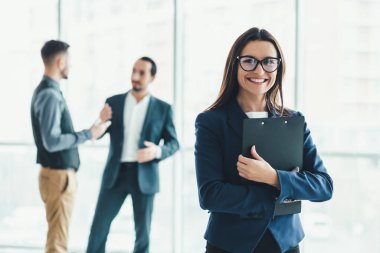 Elinde klasörle güzel bir ofis çalışanının portresi, kameraya gülümsüyor ve meslektaşları bulanık arka planda bir şeyi tartışıyor..