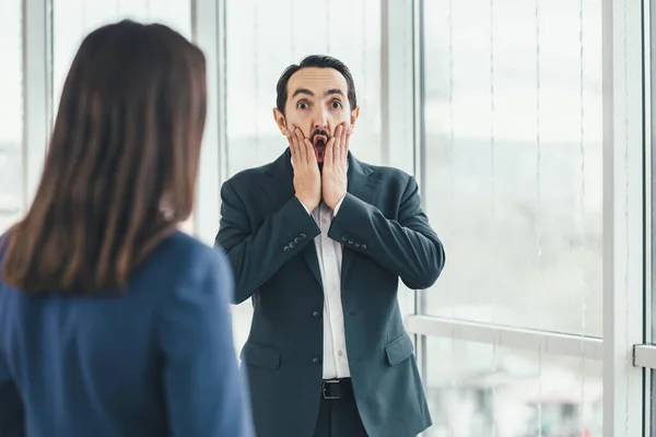 Man looking surprised, open-mouthed, shocked, keeping hands on cheeks, realizing a new thought, idea or some news. Woman is looking at him. — Stock Photo, Image