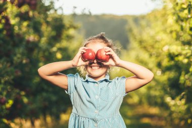 Mavi elbiseli, sevimli, gülümseyen bir çocuk, bulanık arka planda elmalarını gözünün önünde tutuyor..