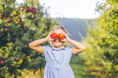 Mavi elbiseli, sevimli, gülümseyen bir çocuk, bulanık arka planda elmalarını gözünün önünde tutuyor..