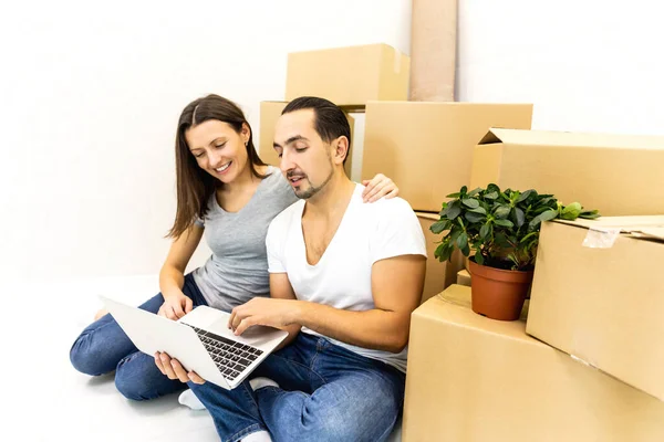 Young family sitting among cardboards, using laptop for surfing in internet to find furniture to their new house. — Stock Photo, Image