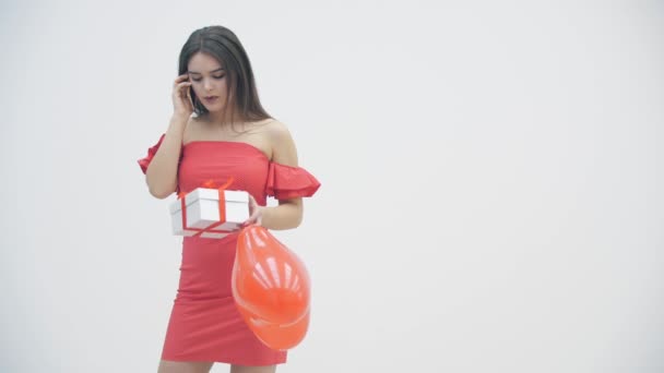 Ragazza delusa con in mano un palloncino rosso e una scatola regalo, mentre parla al telefono con il suo ragazzo che non ha più bisogno dei suoi regali.. — Video Stock
