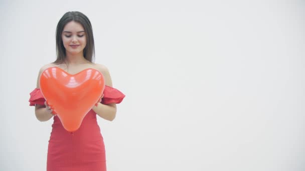 4k video wanita tersenyum berdiri di atas latar belakang putih dengan balon berbentuk hati merah di tangan. — Stok Video
