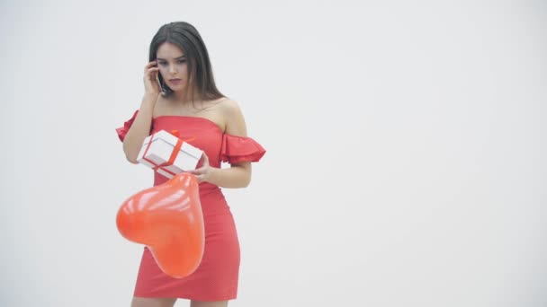 4k video di ragazza delusa con in mano un palloncino rosso e una scatola regalo, mentre parla al telefono con il suo ragazzo che non ha più bisogno dei suoi regali. — Video Stock