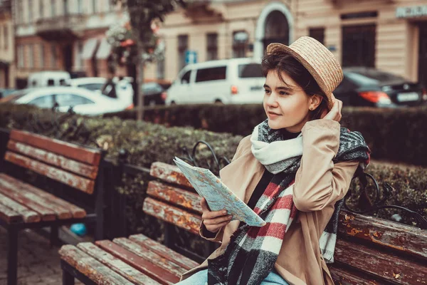 Wanita bersemangat mengunjungi tempat-tempat baru, memegang peta di tangan, mengenakan jas trendi dan topi di atas latar belakang kota yang kabur. — Stok Foto