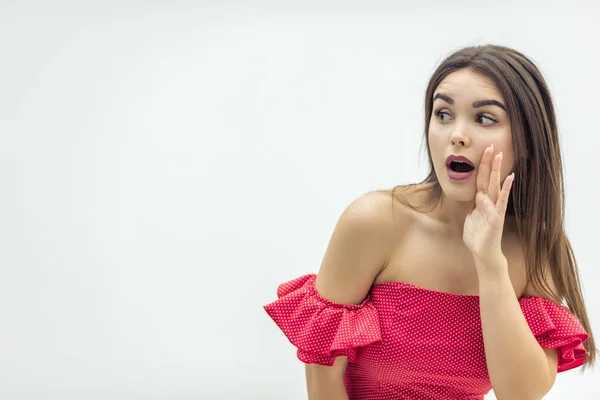 Copyspace foto de la hermosa mujer joven en vestido rojo, chismes, mantener la mano cerca de la boca sobre el fondo blanco . — Foto de Stock