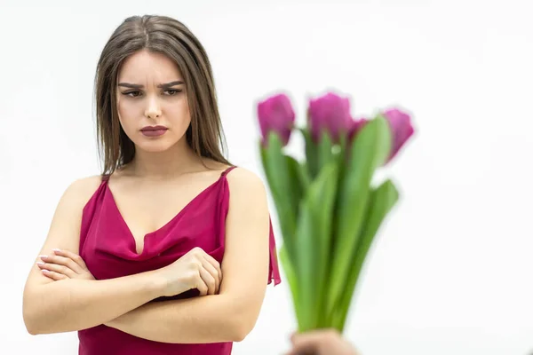 Sad frowning young woman standing with arms crossed and looks displeaased at the tulips man gives her over white background.