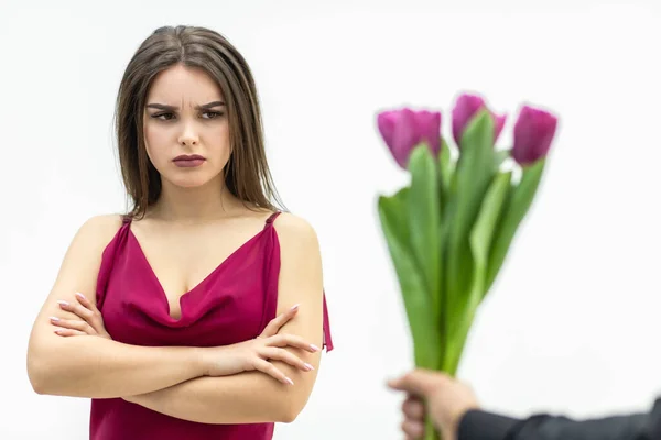 Sad frowning young woman standing with arms crossed and looks displeaased at the tulips man gives her over white background.