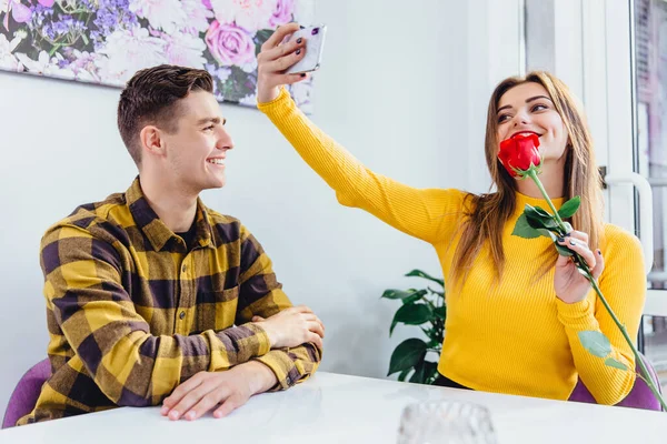 Menina agradável leva selfie com rosa vermelha, que seu namorado lhe deu. Rapaz está realmente feliz por ver tal reacção ao seu presente. . — Fotografia de Stock