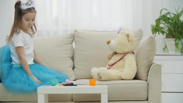 작은 소녀가 최고의 친구 테디 베어와 다과회에서 놀고 있습니다. 소파에 앉아 있죠. 푸른 드레스와 공주의 왕관. — 비디오